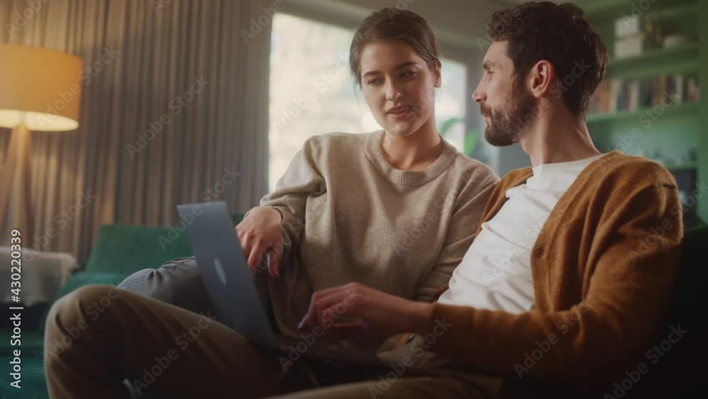 Couple Use Laptop Computer, while Sitting on a Couch in the Apartment. Boyfriend and Girlfriend Talk, Shop on Internet, Choose Product to Order Online, Watch Funny Videos on Streaming Service Couple Use Laptop Computer, while Sitting on a Couch in the Apartment. Boyfriend and Girlfriend Talk, Shop on Internet, Choose Product to Order Online, Watch Funny Videos on Streaming Service