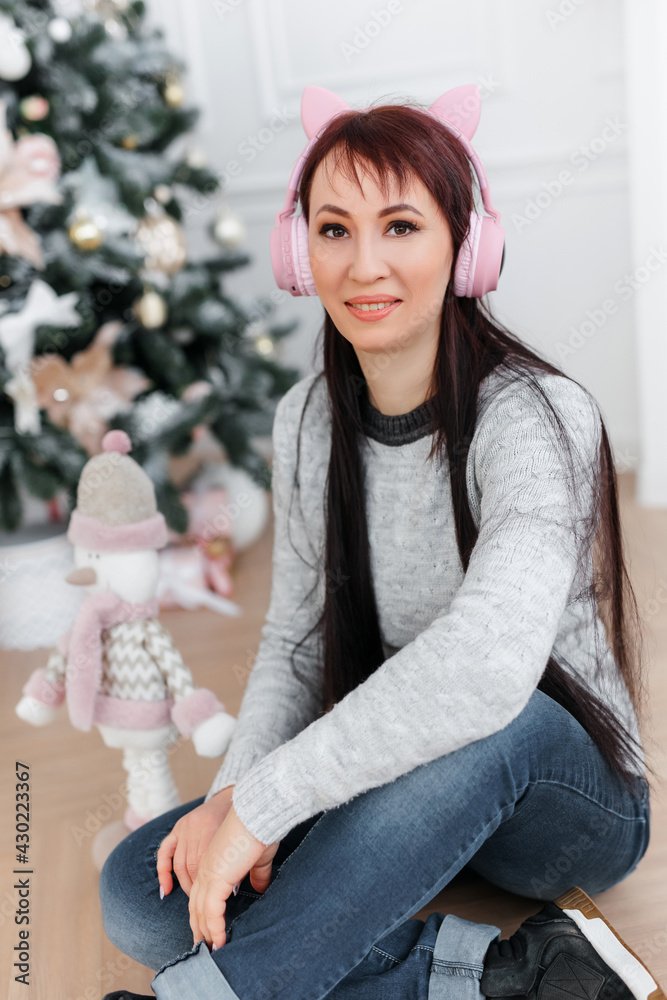 Fototapeta premium caucasian brunette girl with long hair in a knitted sweater. pink headphones with cat ears. the concept of the new year and the holiday. a large artificial Christmas tree with toys in a bright room.