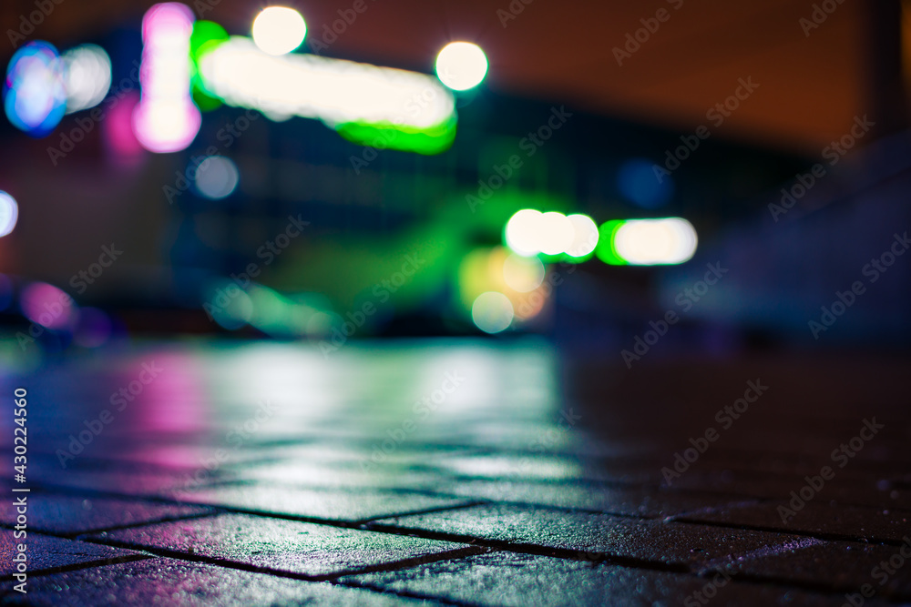 Fototapeta premium Rainy night in the city. Deserted parking mall. Reflections of shop windows on the wet pavement. Colorful colors. Close up view from the pavement level.
