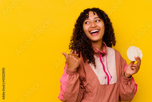 Young mixed race sport woman holding a rice cakes isolated on yellow background points with thumb finger away, laughing and carefree. © Asier