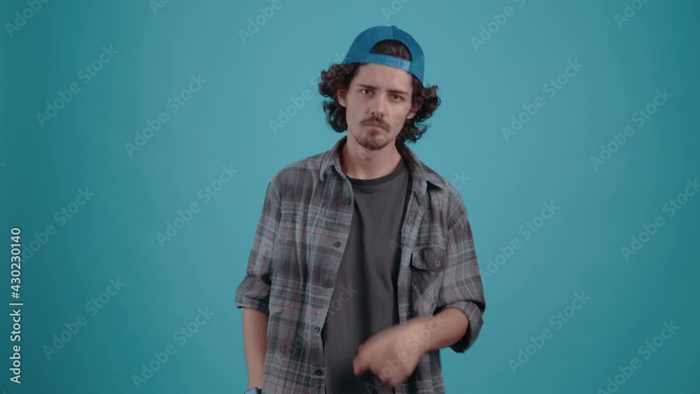 The charismatic young man with the cap, angrily calls someone, with his finger forward and a gesture of coming, puts his fist on his cheek and is ready to beat. isolated over turquoise background The charismatic young man with the cap, angrily calls someone, with his finger forward and a gesture of coming, puts his fist on his cheek and is ready to beat. isolated over turquoise background