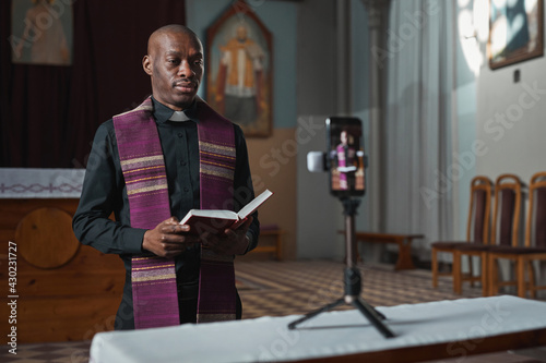 African priest standing in front of the camera on mobile phone and reading the Bible online in the church