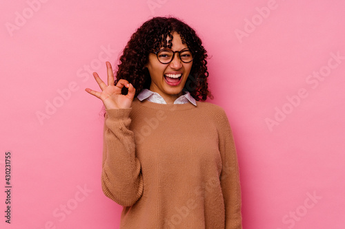 Young mixed race woman isolated on pink background winks an eye and holds an okay gesture with hand. © Asier