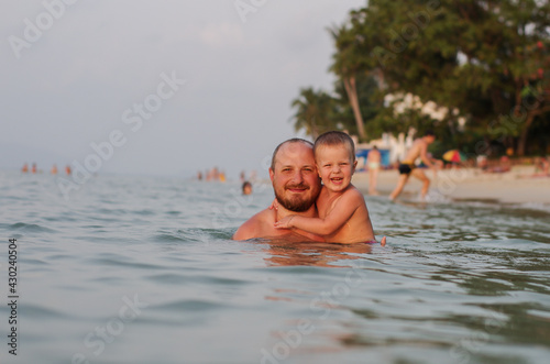 dad and son swim in the sea at sunset. they smile at the camera. the baby and his father spend a happy time on the beach © Julia