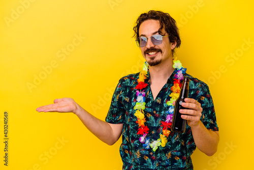 Young caucasian man wearing a hawaiian necklace holding a beer isolated on yellow background showing a copy space on a palm and holding another hand on waist.