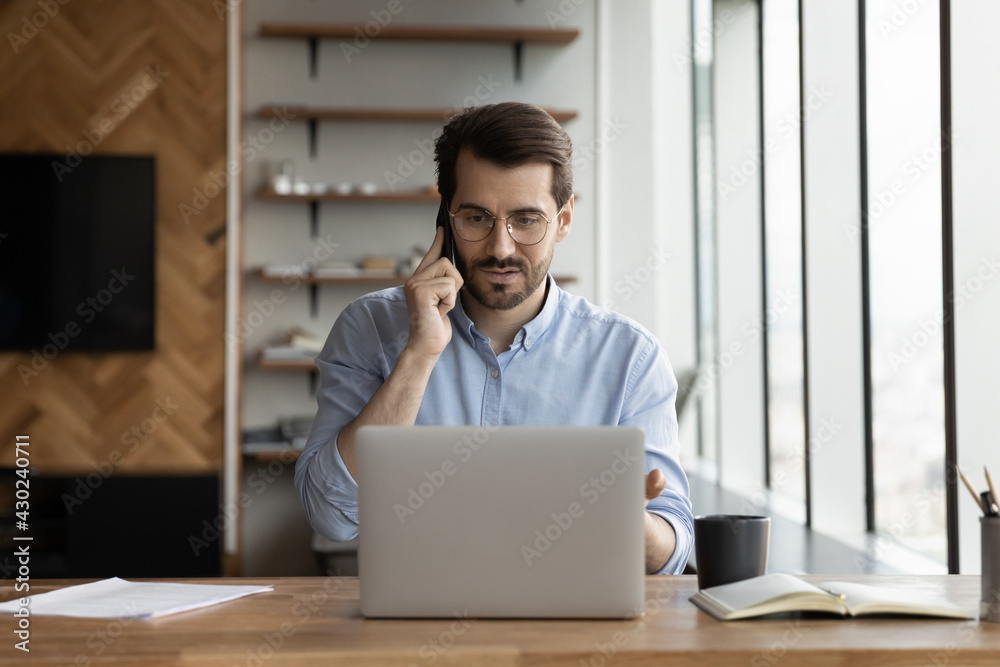 Serious young Caucasian businessman sit at desk at home office work on laptop online talk on smartphone gadget. Confident male employee use computer consult customer client on cellphone call. Serious young Caucasian businessman sit at desk at home office work on laptop online talk on smartphone gadget. Confident male employee use computer consult customer client on cellphone call.