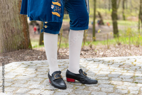 Details of a man's dress in a baroque costume. White stockings and black shoes, golden buttons, decorative hems. A man standing on a cobbled walkway in the background of a blurred garden backdrop.