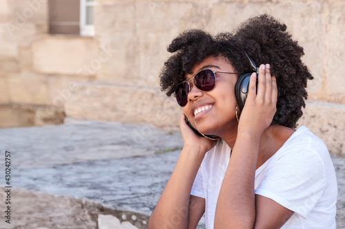 Smiling young black girl with afro hair listening to music with her headphones and sunglasses outdoors on a hot summer day