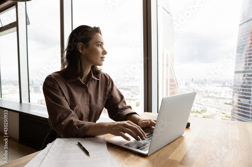 Dreamy young Caucasian businesswoman work on computer in modern office building look in distance making plans of career success or perspectives. Pensive female employee busy with laptop thinking.