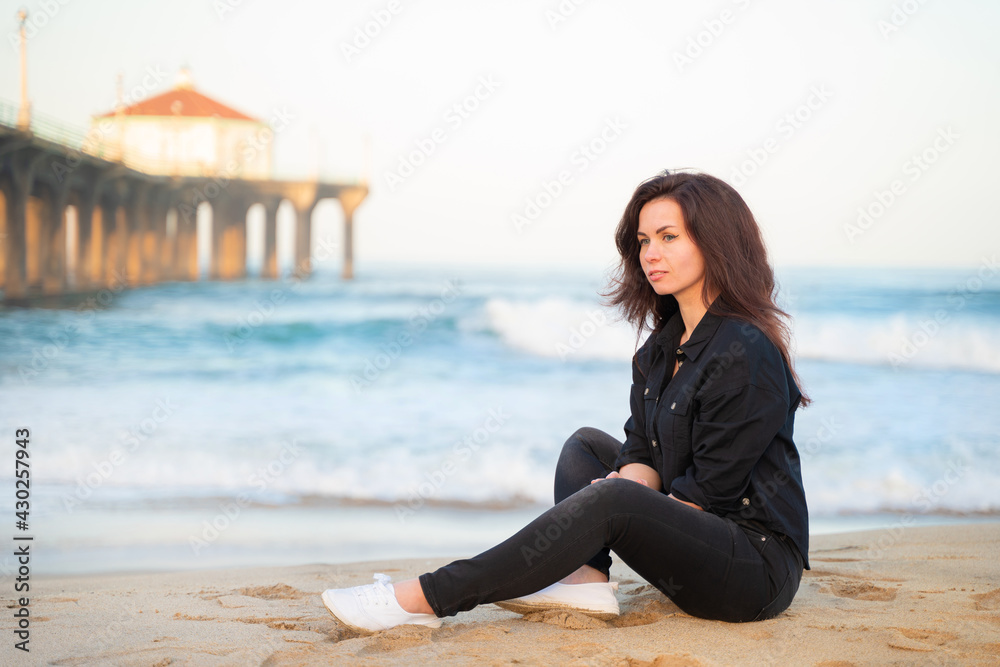 A smiling young woman with long hair sits on the sand in front of a pier on Manhattan Beach in Los Angeles in the early morning
