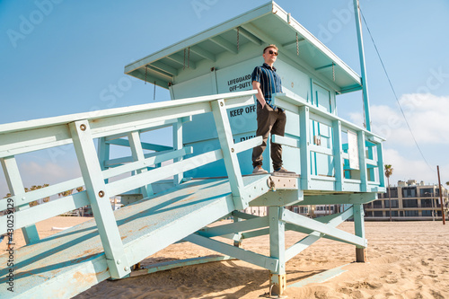 Handsome young blonde man in shirt poses on light blue lifeguard tower on Los Angeles beach