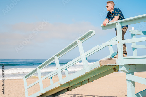 Handsome young blonde man in shirt poses on light blue lifeguard tower on Los Angeles beach