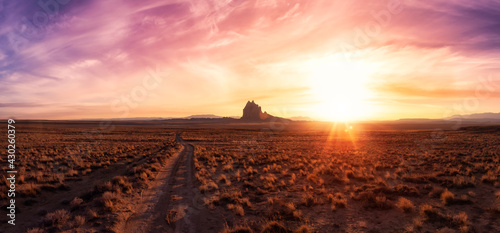 Striking panoramic landscape view of a dirt road in the dry desert with a mountain peak in the background. Colorful Sunset Sky Art Render. Taken at Shiprock, New Mexico, United States.