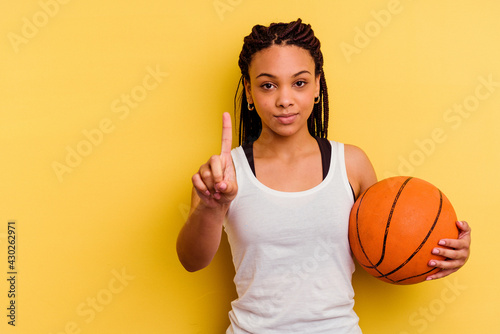 Young african american woman playing basketball isolated on yellow background showing number one with finger. photo