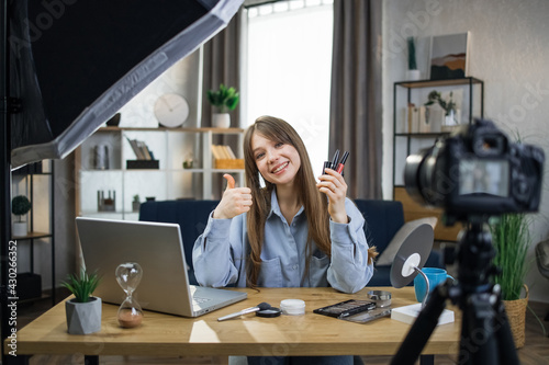 Smiling caucasian woman sitting at table and recording makeup tutorial for sharing in social networks. Beauty blogger testing new cosmetics and showing thumb up.