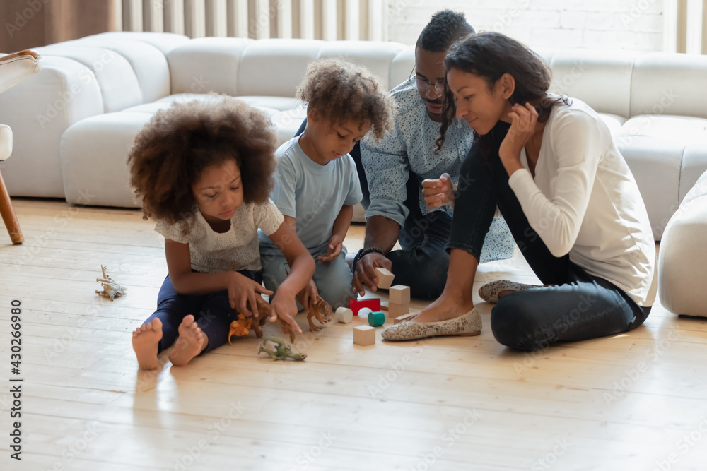 African American parents and two preschooler kids playing toy blocks and dinosaurs on heat floor in living room. Happy family enjoying weekend leisure time at home. Couple engaged in children playtime African American parents and two preschooler kids playing toy blocks and dinosaurs on heat floor in living room. Happy family enjoying weekend leisure time at home. Couple engaged in children playtime