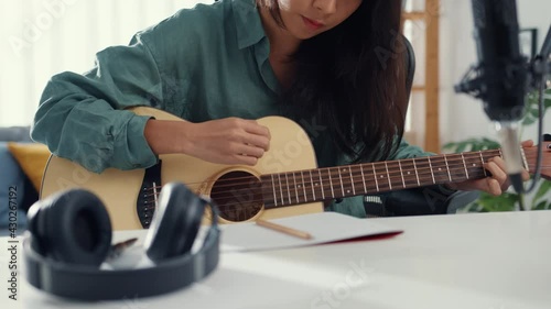 Happy asia woman songwriter play acoustic guitar listen song from smartphone think and write notes lyrics song in paper sit in living room at home studio. Music production at home concept. photo