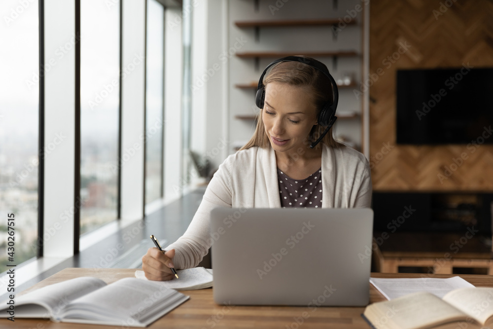 Focused young Caucasian woman in headphones write take notes study learn distant on computer at home. Millennial female in earphones handwrite talk on webcam video call, have zoom class on laptop.