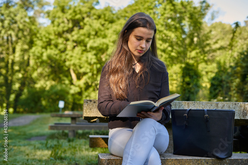 long-haired girl, black shirt and white pants reading a book in nature, sitting at a stone table with a coffee and her bag. reading with background trees and green book teenager studying in the park