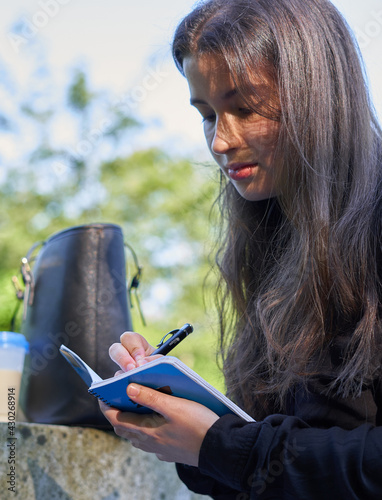 long-haired pretty woman sitting on a stone table in nature writing in a notebook with coffee and her bag. teenager drawing in a blue notebook and black pen in the forest. girl making notes in agenda