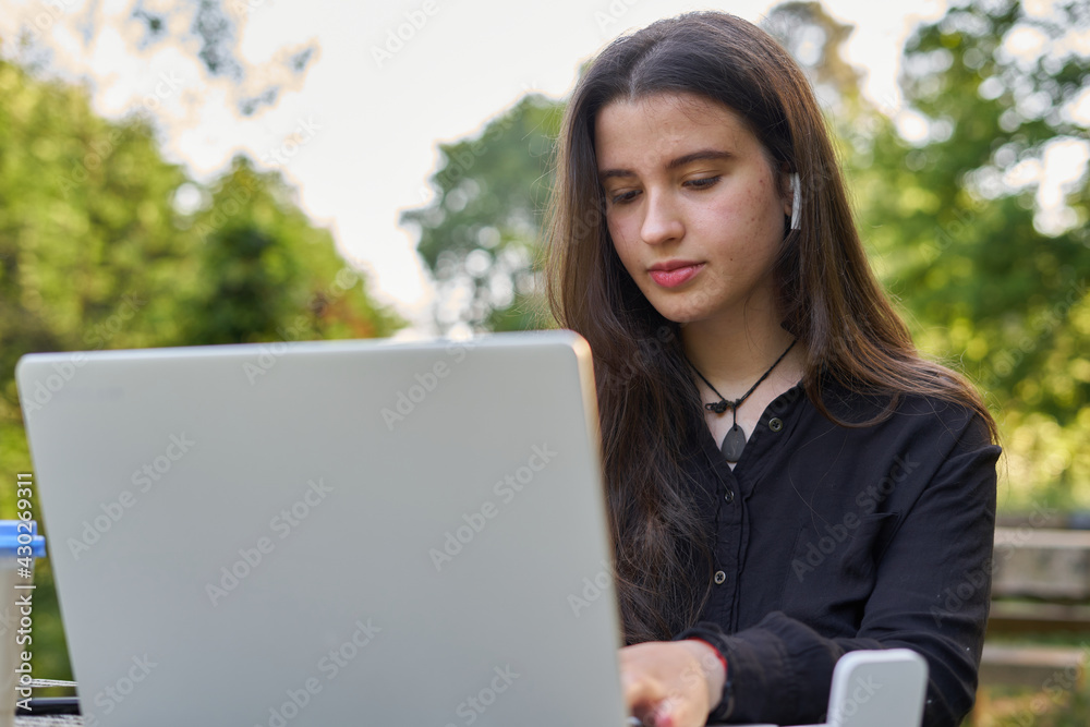 long-haired pretty businesswoman with laptop, headphones and coffee at stone table. teenager working on her laptop in nature with unfocused green background. girl with computer outdoors. long-haired pretty businesswoman with laptop, headphones and coffee at stone table. teenager working on her laptop in nature with unfocused green background. girl with computer outdoors.
