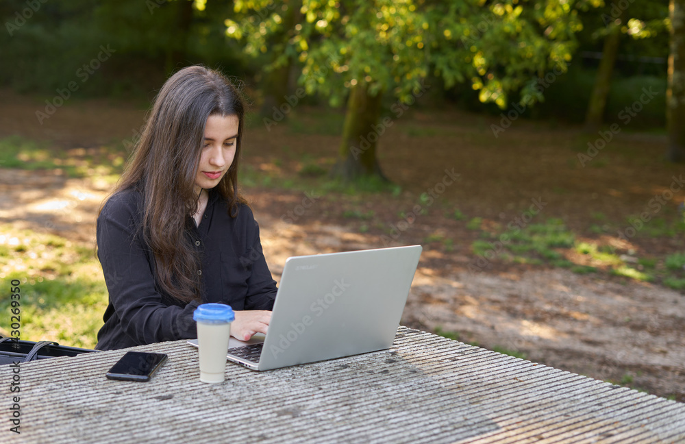 long-haired pretty businesswoman with laptop, headphones and coffee at stone table. teenager working on her laptop in nature with unfocused green background. girl with computer outdoors. long-haired pretty businesswoman with laptop, headphones and coffee at stone table. teenager working on her laptop in nature with unfocused green background. girl with computer outdoors.