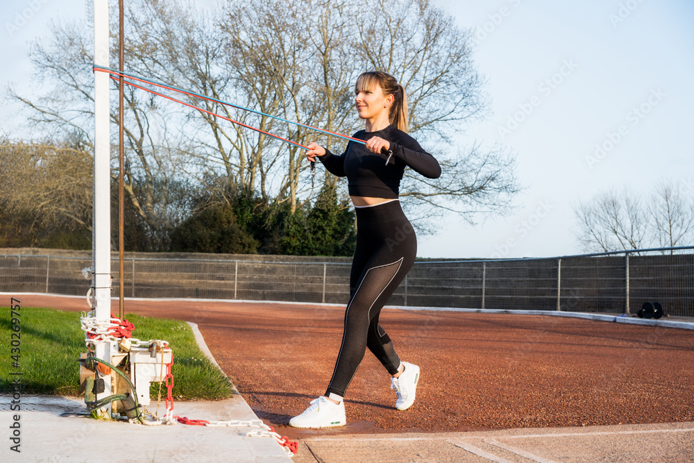 Young fitness woman in sportswear doing standing exercise for thorax muscles with resistance band during outdoors workout on the stadium. Active lifestyle, sport and health care. Copy space