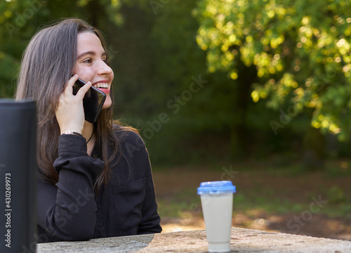 long-haired, fair-skinned woman sitting at a stone table with a coffee and her bag while talking on the phone, with green unfocused background. teenager in nature with his mobil taking a break.