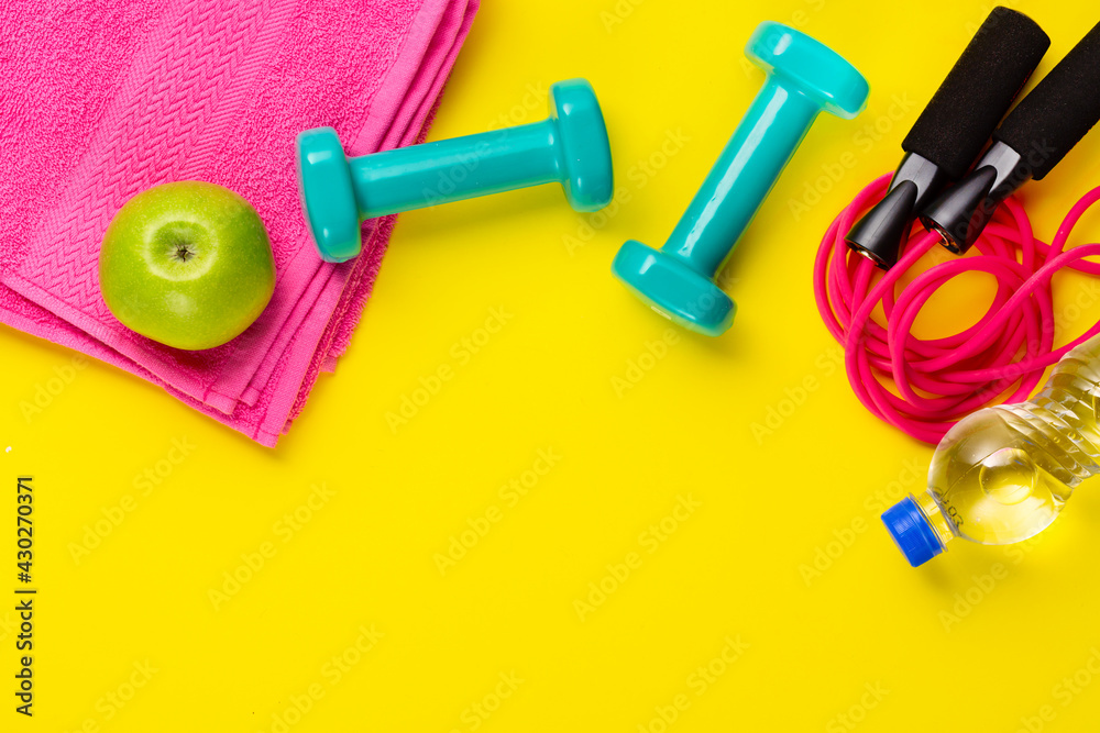Fitness and sport concept. Skipping rope, green apple, dumbbells, bottle of water and pink towel on bright yellow background. Free space Fitness and sport concept. Skipping rope, green apple, dumbbells, bottle of water and pink towel on bright yellow background. Free space