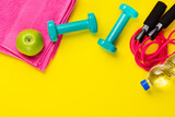 Fitness and sport concept. Skipping rope, green apple, dumbbells, bottle of water and pink towel on bright yellow background. Free space