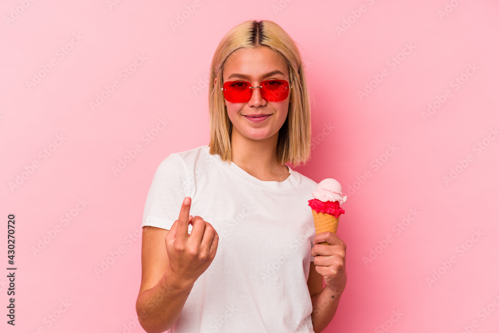 Young venezuelan woman eating an ice cream isolated on pink background pointing with finger at you as if inviting come closer. Young venezuelan woman eating an ice cream isolated on pink background pointing with finger at you as if inviting come closer.