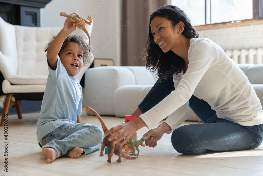 Fototapeta premium Happy cute kid boy and his mom relaxing on heat floor in cozy living room and playing with small dinosaurs figures together. Mother engaged in little sons game with toys. Family playtime concept