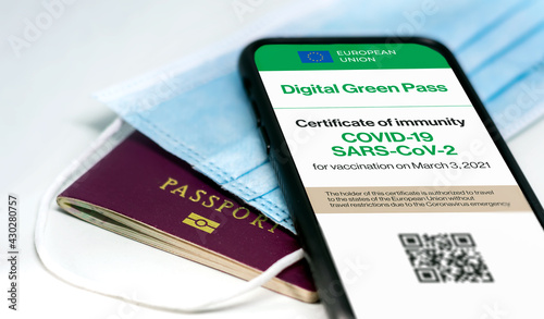 The digital green pass of the european union with the QR code on the screen of a mobile phone over a surgical mask and a passport