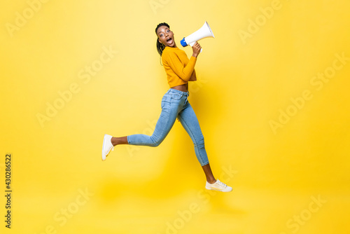 Jumping portrait of shocked young African American woman holding megaphone making announcement in isolated studio yellow background