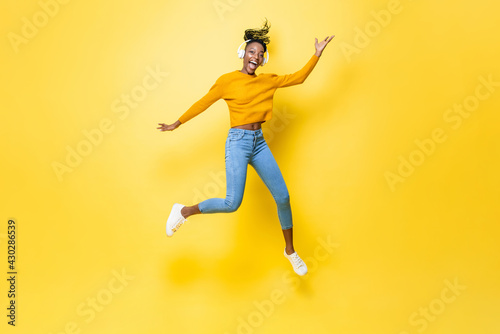 Happy energetic young African American woman wearing headphones listening to music and jumping with hand up in yellow isolated studio background