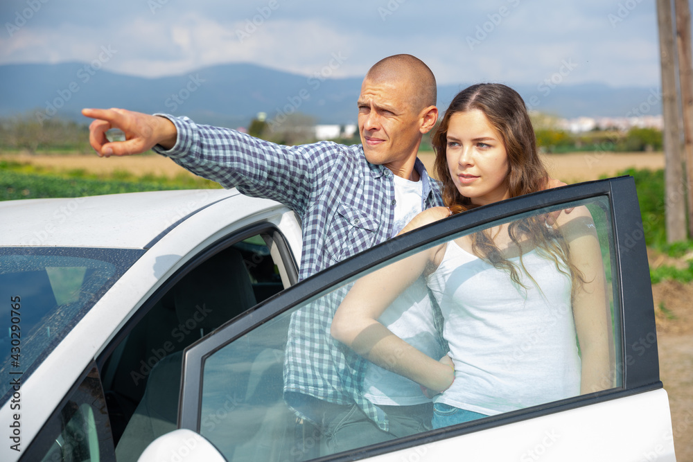 Positive young woman and man standing near car and pointing to something