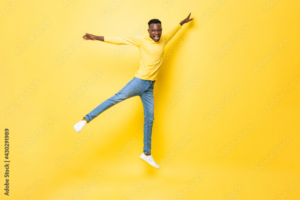 Young happy energetic African man jumping with open arms and legs on isolated yellow studio background