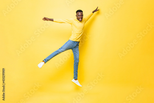 Young happy energetic African man jumping with open arms and legs on isolated yellow studio background