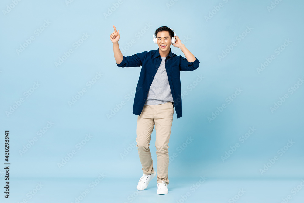 Full body portrait of happy smiling young handsome Asian man listening to music with wireless headphones pointing hand up on isolated light blue studio background Full body portrait of happy smiling young handsome Asian man listening to music with wireless headphones pointing hand up on isolated light blue studio background