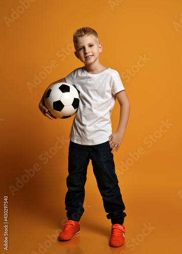 Fototapeta Naklejka Na Ścianę i Meble -  Happy smiling schoolboy, teenager, soccer fan in casual clothes with white blank t-shirt stands holding soccer ball in hand over yellow background