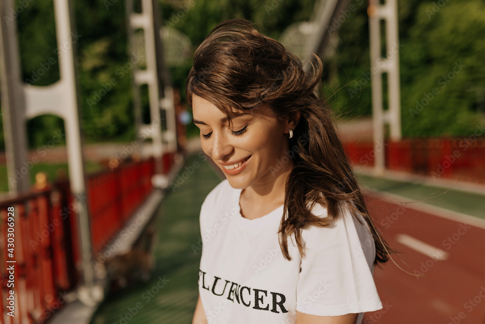 Charming adorable lady with long hair wearing white t-shirt smiling with closed eyes in sunlight in the morning Charming adorable lady with long hair wearing white t-shirt smiling with closed eyes in sunlight in the morning