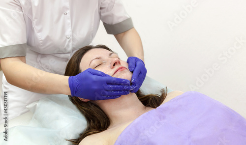 Cosmetologist hands in blue gloves touch face of caucasian woman patient making massage procedure in beauty salon.