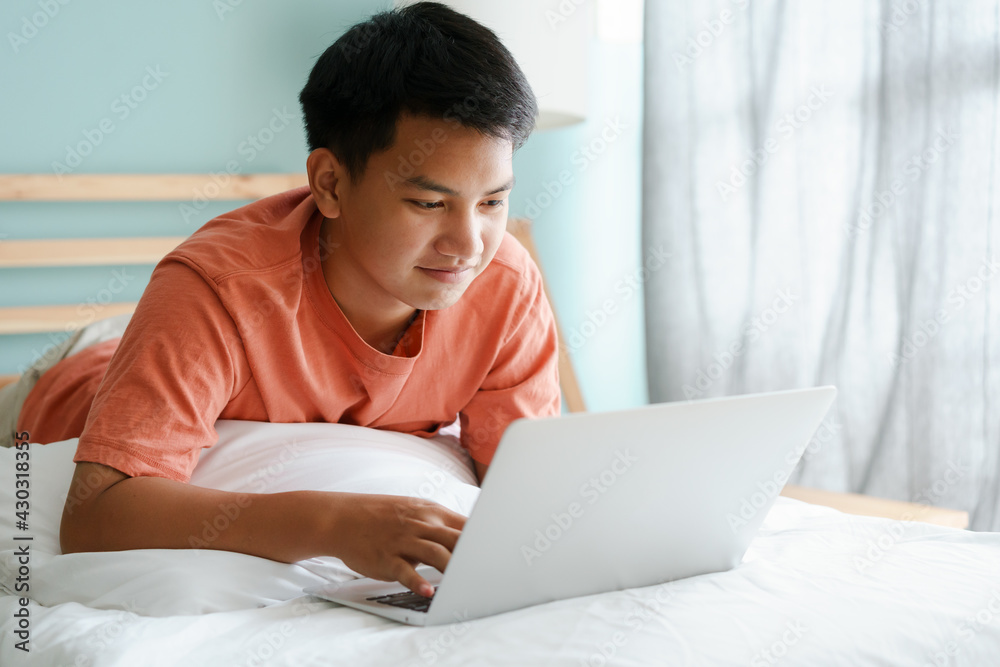 Asian man using a computer laptop while lying on a bed in the morning Asian man using a computer laptop while lying on a bed in the morning