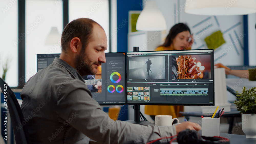 Video maker editing movie using post production software working in creative start up agency office with two monitors. Videographer processing audio film montage on professional computer, taking notes Video maker editing movie using post production software working in creative start up agency office with two monitors. Videographer processing audio film montage on professional computer, taking notes