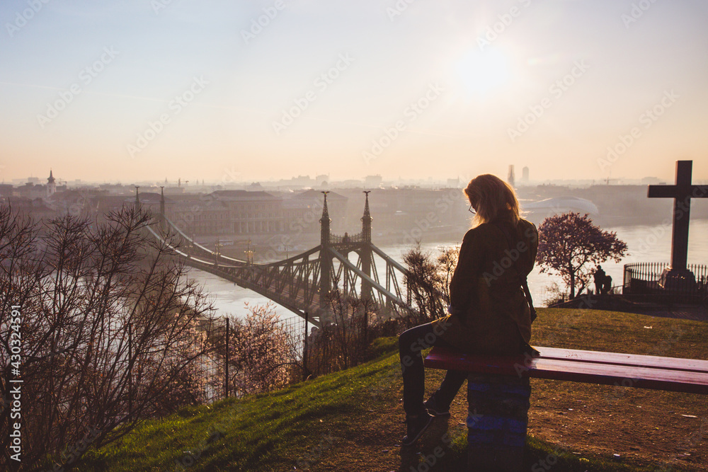 Fototapeta premium Woman sitting on the bench and looking to the Liberty bridge over Danube from the terrace of the Gellert hill in Budapest, Hungary on sunrise