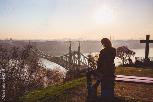 Woman sitting on the bench and looking to the Liberty bridge over Danube from the terrace of the Gellert hill in Budapest, Hungary on sunrise