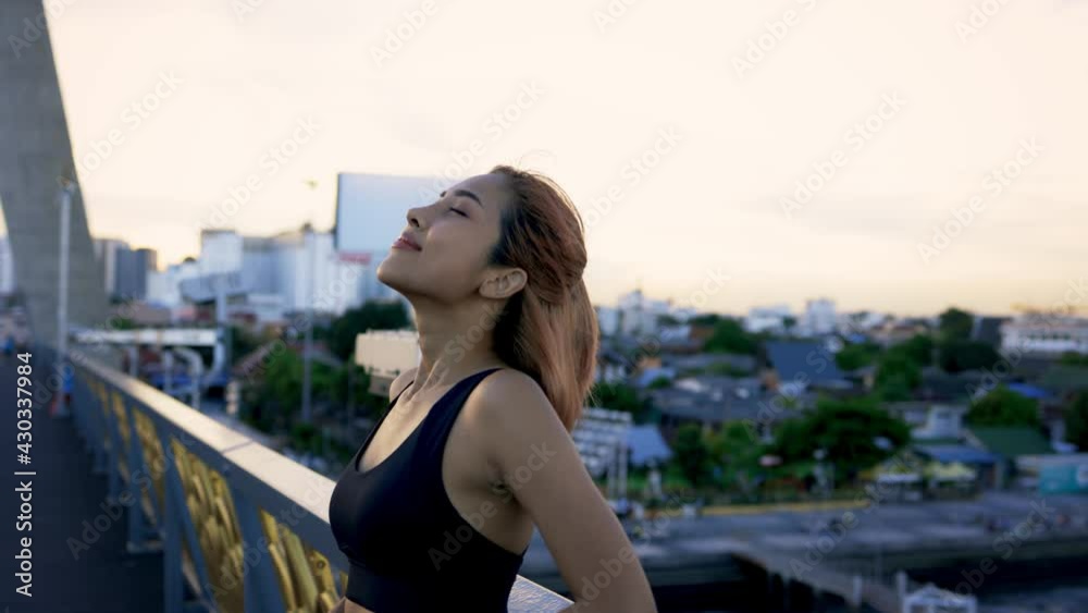 Healthy and fit young Asian female athlete leaning on bridge railing taking fresh deep breathe for relaxation after running in evening Healthy and fit young Asian female athlete leaning on bridge railing taking fresh deep breathe for relaxation after running in evening