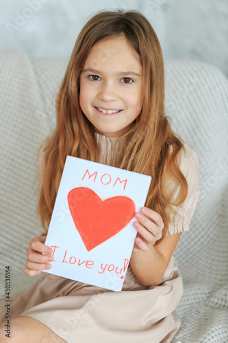 Laughing child girl holding greeting homemade drawing card red heart. Gift for Mothers Day. Happy childhood concept. Inscription Mom, I love you. Attractive daughter congratulate mum, happy birthday