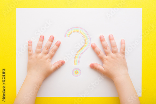 Baby hands and one colorful question sign drawn on white paper on bright yellow table background. Closeup. Point of view shot. Children issues. Top down view.