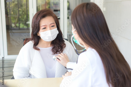 Asian woman doctor uses stethoscope to check health of lungs and heart or diagnosis symptom of woman patient while both wear face mask at hospital in health care and coronavirus protection concept.